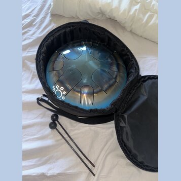 Modrý kovový steel tongue drum umístěný v černém polstrovaném přenosném pouzdře. Na boku nástroje jsou bílé nápisy G, G, F, A, O, D. Vedle pouzdra le…