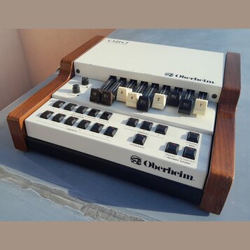 Bílý elektronický klávesový syntetizátor s dřevěnými bočními panely. Na horním panelu je nápis OB-3 a logo Oberheim, v dolní části je opět logo Oberh…