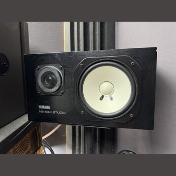 Černý studiový monitor s dřevěným povrchem. Na přední straně je bílý basový reproduktor a menší výšek s kovovou mřížkou. V levém dolním rohu je bílý…