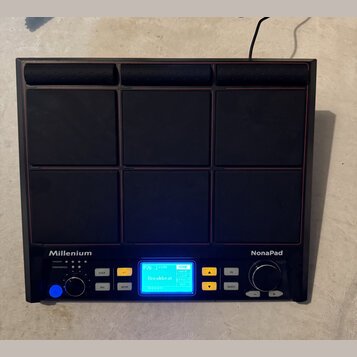Černý elektronický perkusní pad s devítitlačidlovým rozložením 3x3. Na spodním panelu jsou bílé nápisy Millenium a NonaPad, uprostřed se nachází modř…