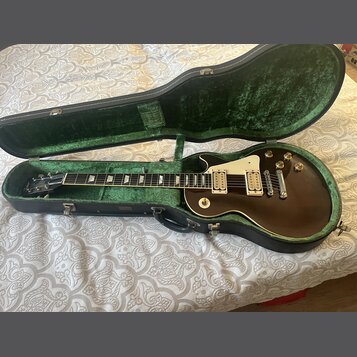 Tmavně hnědá elektrická kytara typu Les Paul s krémovým pickguardem a dvěma humbuckery v černém tvrdém kufru s zeleným polstrováním. Na hmatníku jsou…