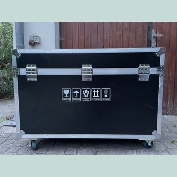 Černý transportní flightcase s hliníkovými hranami a kovovými rohy. Přední strana obsahuje tři stříbrné zapínací zámky a bílé informační piktogramy s…