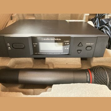 Černý bezdrátový přijímač s nápisem audio-technica. a označením UHF SYNTHESIZED DIVERSITY RECEIVER ATW-R3100. Obsahuje displej, tlačítka POWER a MODE…