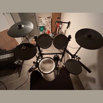 Elektronická bicí souprava s černými pady a činely na kovovém stojanu. Na bílém snare padu je vidět nápis Roland. Soubor obsahuje pět padů a dva čine…