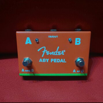 Oranžový efekt pedál s nápisem Fender a označením ABY PEDAL. Na horní části jsou dva vstupy/výstupy označené písmeny A a B a nápis IN/OUT. V dolní čá…