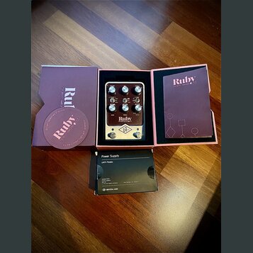 Kytarový pedál v barvě stříbrné a černé, umístěný v originální fialové krabici. Na pedálu je nápis Ruby, TOP BOOST AMPLIFIER '63 a logo UA v kosočtve…
