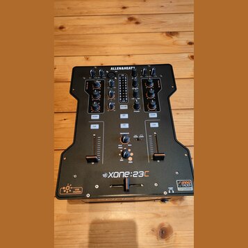 Černý DJ mixér s nápisem ALLEN&HEATH v horní části a XONE:23C ve spodní části. Zařízení obsahuje několik faderů, otočných knoflíků a tlačítek s popis…