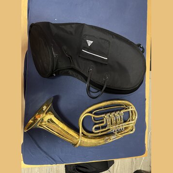Na fotografii je mosazný železný nástroj typu baryton nebo euphonium v zlaté barvě a černý polotvrdý pouzdro. Na pouzdře je vidět bílé trojúhelníkové…