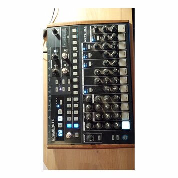 Černý analogový syntezátor s dřevěným rámem. Na zařízení je nápis DRUMBRUTE ANALOG DRUM SYNTHESIZER a logo ARTURIA s textem YOUR EXPERIENCE SOUND. Př…