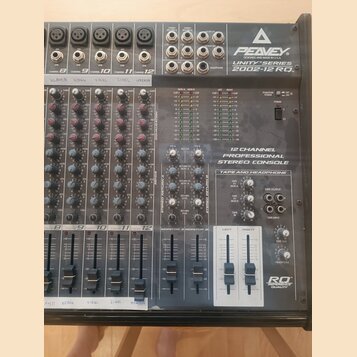 Šedý 12kanálový mixážní pult s nápisy PEAVEY, UNITY SERIES a 2002-12 RQ. Na přístroji je text 12 CHANNEL PROFESSIONAL STEREO CONSOLE a v pravém dolní…