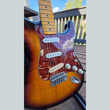 Elektrická kytara s trupem v barevném přechodu od žluté přes oranžovou k hnědé. Nástroj má hnedý mramorovaný pickguard, tři bílé snímače a javorový h…