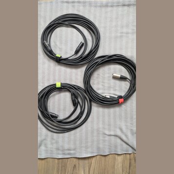 Tři srolovaný černé audio kabely. Dva z nich mají koncovky typu XLR, jeden má jednu koncovku typu jack. Kabely jsou stahovány barevnými suchými staho…