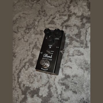 Černý efekt pedál s nápisy Blond a DISTORTION. Na horní straně jsou tři ovládací knoflíky s popisky G, LEVEL a TONE, přepínač HIGH/LOW a vstup pro na…
