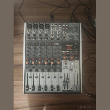 Šedý mixovací pult značky behringer s nápisem 24-BIT MULTI-FX PROCESSOR. Zařízení obsahuje několik vstupních kanálů s posuvníky, červené a modré ovlá…