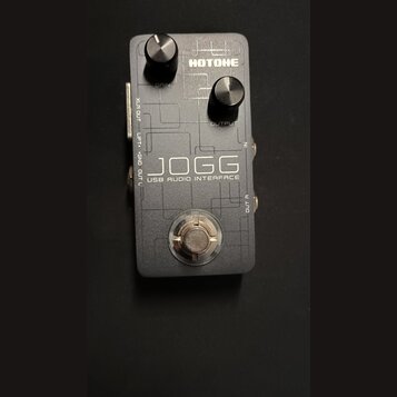 Šedý obdélníkový USB audio interface s kovovým povrchem. Na horní straně jsou dva černé ovládací knoflíky s popisky GAIN a OUTPUT a logo HOTONE. Upro…
