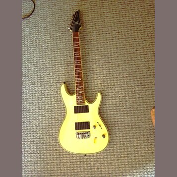 Žlutá elektrická kytara s tmavým hnedým hmatníkem a černou hlavou. Na hlavě je viditelné logo značky Ibanez. Nástroj má dva černá humbuckery, jeden o…