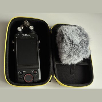 Černý digitální záznamový zařízení v žlutém přepravním pouzdře. Na horní části přístroje je bílý nápis TASCAM a pod ním Portacapture X8. V pravé část…