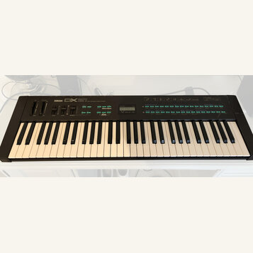 Černý digitální klavír s bílými a černými klávesami. Na horním panelu je nápis YAMAHA DX21, malý displej a řada zelených tlačítek. Nástroj vykazuje m…