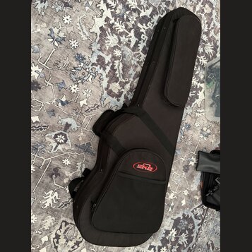Černý polotvrdý pouzdro (gig bag) na hudební nástroj s dlouhým úzkým tvarem. Na přední kapsi je umístěno červenobílé logo SKB. Pouzdro má připevněný…