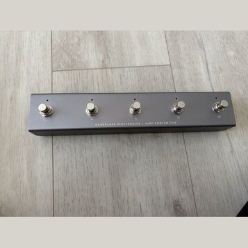 Šedý MIDI footswitch v kovovém obalu s pěti stříbrnými spínači. Na spodní hraně je bílý nápis DARKGLASS ELECTRONICS - MIDI FOOTSWITCH. Povrch zařízen…