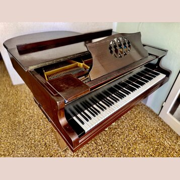 Hnědé dřevěné piano s otevřeným horním víkem, skrze které jsou vidět kovové struny a vnitřní mechanismus. Na hudebním pultu je vyřezaný kruhový ornam…