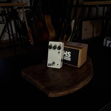 Bílý efekt pedál s nápisem FUZZ a logem JHS SERIES. Na horní straně jsou tři ovládací knoflíky s popisky VOLUME, BASS a FUZZ. Vedle pedálu je umístěn…
