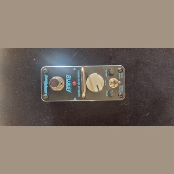 Kytarový efekt pedál v kovovém stříbrném provedení. Na povrchu jsou nápisy „TomMoye“, „BLUESY“ a „VINTAGE BLUES OVERDRIVE“. Pedál má jeden velký bílý…