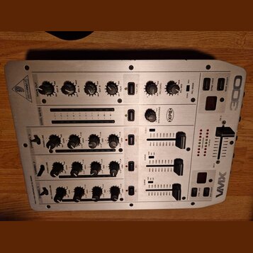 Stříbrný DJ mixér s nápisy BEHRINGER a VMX 100. Na horní části jsou ovládací prvky pro MAIN OUT, OUT A a OUT B, vpravo je vidět logo XPO. Přístroj ob…