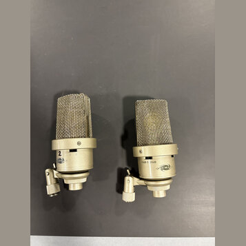 Dva kondenzátorové mikrofony stříbrné barvy s kovovými mřížkami. Na těle pravého mikrofonu je nápis -10dB 0 20dB a logo NEUMANN. Levý mikrofon má na…