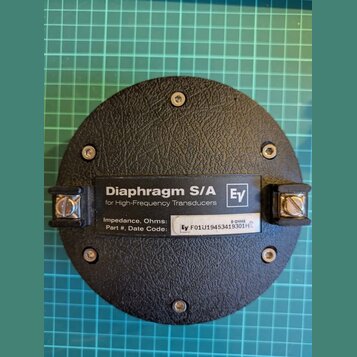 Černý kulatý komponent s texturovaným povrchem, připevněný čtyřmi šrouby. Uprostřed je umístěn štítek s nápisy „Diaphragm S/A“, „for High-Frequency T…