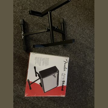 Černý kovový stojan na zesilovač s logem Fender na jedné z nožních gumových konцоvek. Vedle stojanu je umístěna originální krabice s nápisem Fender A…