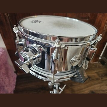 Stříbrný kovový snare drum s bílými koženem. Na korpusu je viditelný oválný štítek s nápisy MAPEX, MPX a STEEL. Nástroj vykazuje mírné znečištění pov…