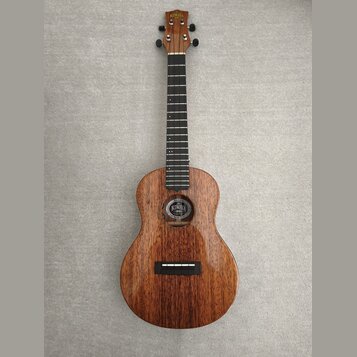Hnědý dřevěný ukulele s tmavým hmatníkem a černými ladičkami. Uvnitř ozvučnice je vidět štítek s nápisem "KALA". Na hlavě nástroje je bílý nápis "KAL…