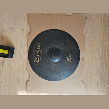 Kruhový činely z tmavého kovaného kovu s viditelnou strukturou kladivování. Na povrchu je žlutý nápis „Turkish“ a pod ním menší text „Handmade cymbal…