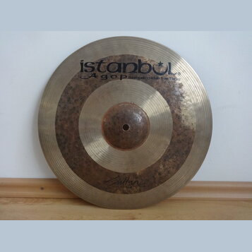 Kovový činel zlatavé barvy s viditelným Oxidováním a tmavými skvrnami v prostřední části. V horní části je černý nápis istanbul Agop Handmade cymbals…