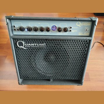 Černý basový kombozesilovač s kovovou mřížkou reproduktoru. Na předním panelu je nápis HUGHES & KETTNER QUANTUM BASS TECHNOLOGY a označení modelu QC…