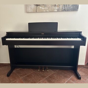 Černá digitální pianola s klaviaturou a stojanem. Na horní části je v centru bílý nápis YAMAHA. Nástroj obsahuje tři pedály a na horní desce je umíst…