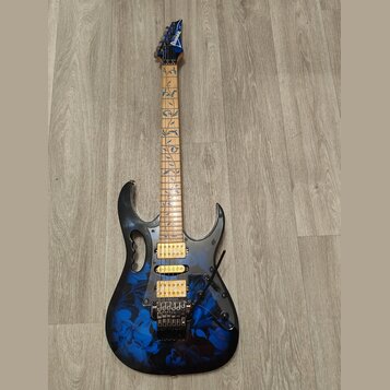 Elektrická kytara s modře a černě strakatým povrchem korpusu a hnědým hmatníkem s modrými ornamenty listů. Na hlavě je bílý nápis Ibanez. Nástroj má…
