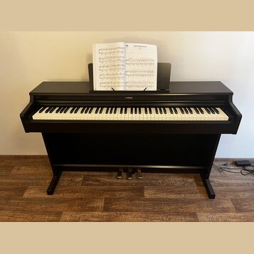 Černý digitální piano s bílými a černými klávesami. Nad klaviaturou je umístěno logo YAMAHA a na notovém pultu jsou položeny notové listy. Nástroj st…