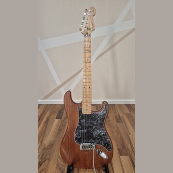 Elektrická kytara s hnědým dřevěným korpusem a světle hnědým javorovým hmatníkem. Na hlavě je viditelné logo Fender. Kytara má černý mramorovaný pick…