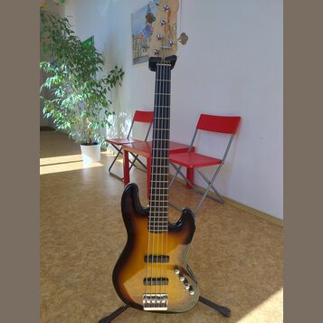 Elektrická basová kytara s pěti strunami a korpusem v barevném přechodu od žluté přes oranžovou k hnědé (sunburst). Na hlavě hmatníku je viditelný ná…