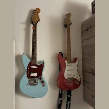 Dvě elektrické kytary zavěšené na bílé stěně. Levá kytara je světle modrá s tmavě červeným pickguardem a na hlavě má nápis Fender. Pravá kytara je če…