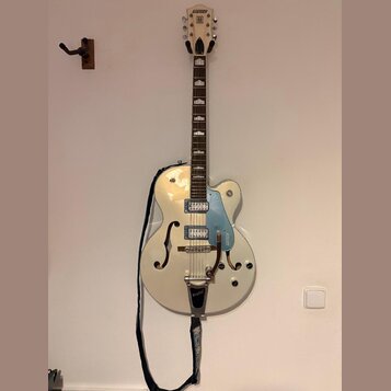 Bílá polodupatá elektrická kytara s modrým pickguardem a chromovaným hardwarem. Na hlavě hmatníku je nápis GRETSCH a na pickguardu je svisle umístěný…