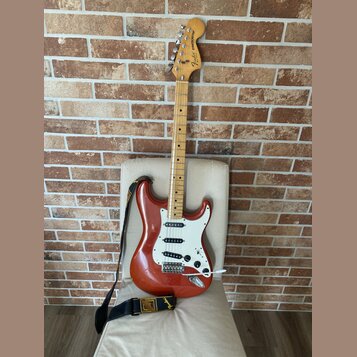Elektrická kytara typu Stratocaster v červeně hnědé barvě s bílým pickguardem a světle hnědým hnedobým hmatníkem. Na hlavě je viditelné logo Fender S…