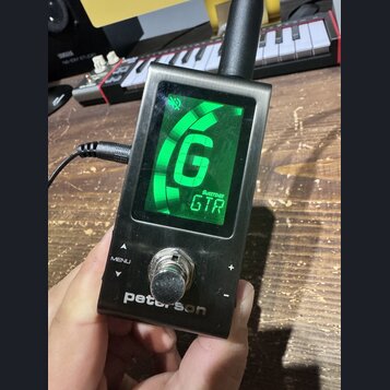 Kompaktní digitální ladič stříbrné barvy s displejem. Na spodní části je bílý nápis peterson a nad ním ovládací prvky MENU, +, - a kovové kolečko. Na…