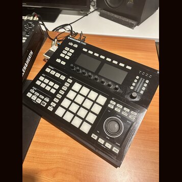 Černý kontrolér Maschine Studio s mřížkou 16 bílých podsvícených padů. Na horní hraně je nápis MASCHINE STUDIO s logem výrobce. Zařízení obsahuje dvě…