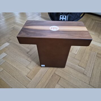 Hnědý dřevěný cajón s logem MEINL v bílém kruhu na horní hraně. Nástroj má kompaktní tvar s vyčnívající horní deskou a užším spodním tělem. Povrch dř…