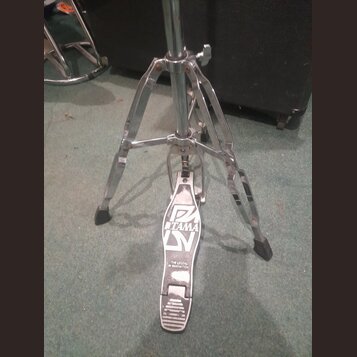 Chromovaný kovový stojan s pedálem, pravděpodobně hi-hat. Na pedálu je viditelné logo TAMA a nápis THE LEGEND IN INNOVATION. Povrch pedálu vykazuje z…