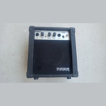 Černý malý kytarový kombo zesilovač s bílým ovládacím panelem. Na panelu jsou nápisy REEF AUDIO, LA5, INPUT, GAIN, VOLUME, TONE, PHONES a DC9V. Na sp…