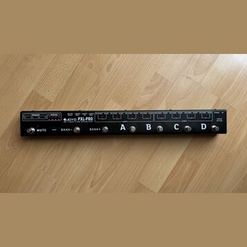 Černý pedálboard s nápisy JOYO PXL-PRO a DOUB 4 CHANNEL FX PEDAL SWITCHING SYSTEM. Zařízení obsahuje čtyři stomečovací spínače označené písmeny A, B,…